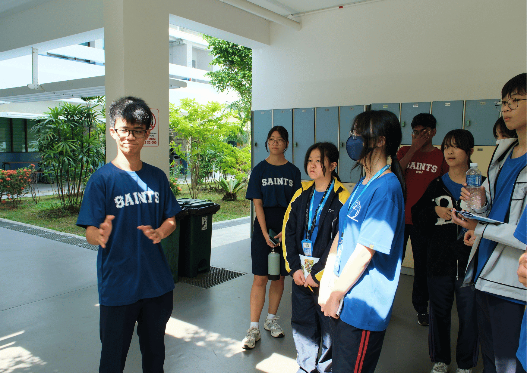 A student from St Andrew's Junior College in a navy "Saints" T-shirt speaks and gestures to a small group of visiting students in blue T-shirts near a row of lockers, appearing to lead a campus tour.